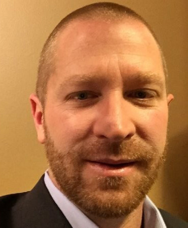 Professional headshot of Dan Wenger. He has a reddish beard and short hair, and is wearing a navy jacket over a light blue dress shirt. He is posed in front of a beige background and is slightly smiling.