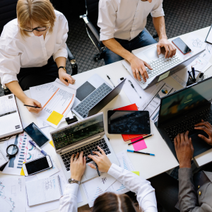 Group of people sitting at a table typing on laptops with papers, phones, and office supplies spread across the table.