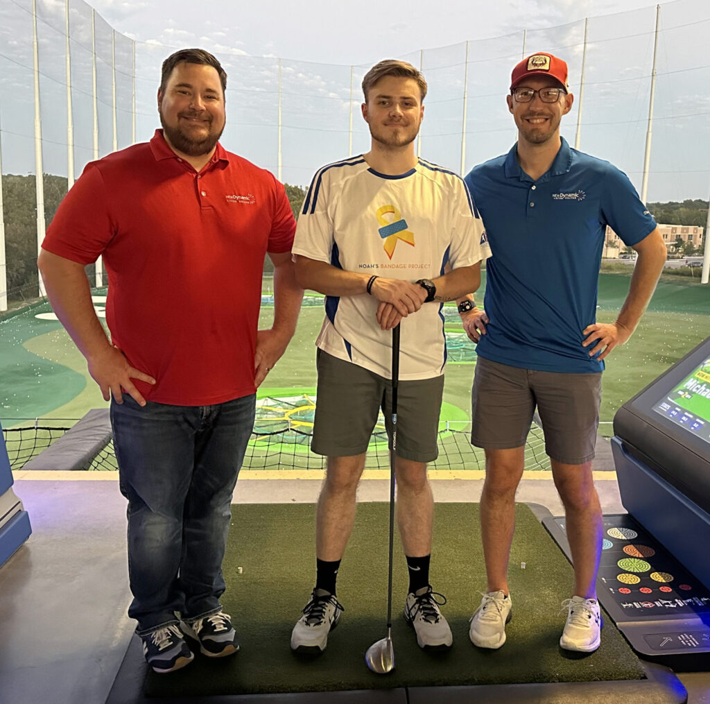 Members of the New Dynamic team at the Tee Off Against Childhood Cancer event. There are three men standing in front of a driving range, the man on the left is in a red shirt with his hands on his hips, the man in the middle is wearing a white shirt and is holding a golf club, and the man on the right is in a blue shirt with his hands on his hips.