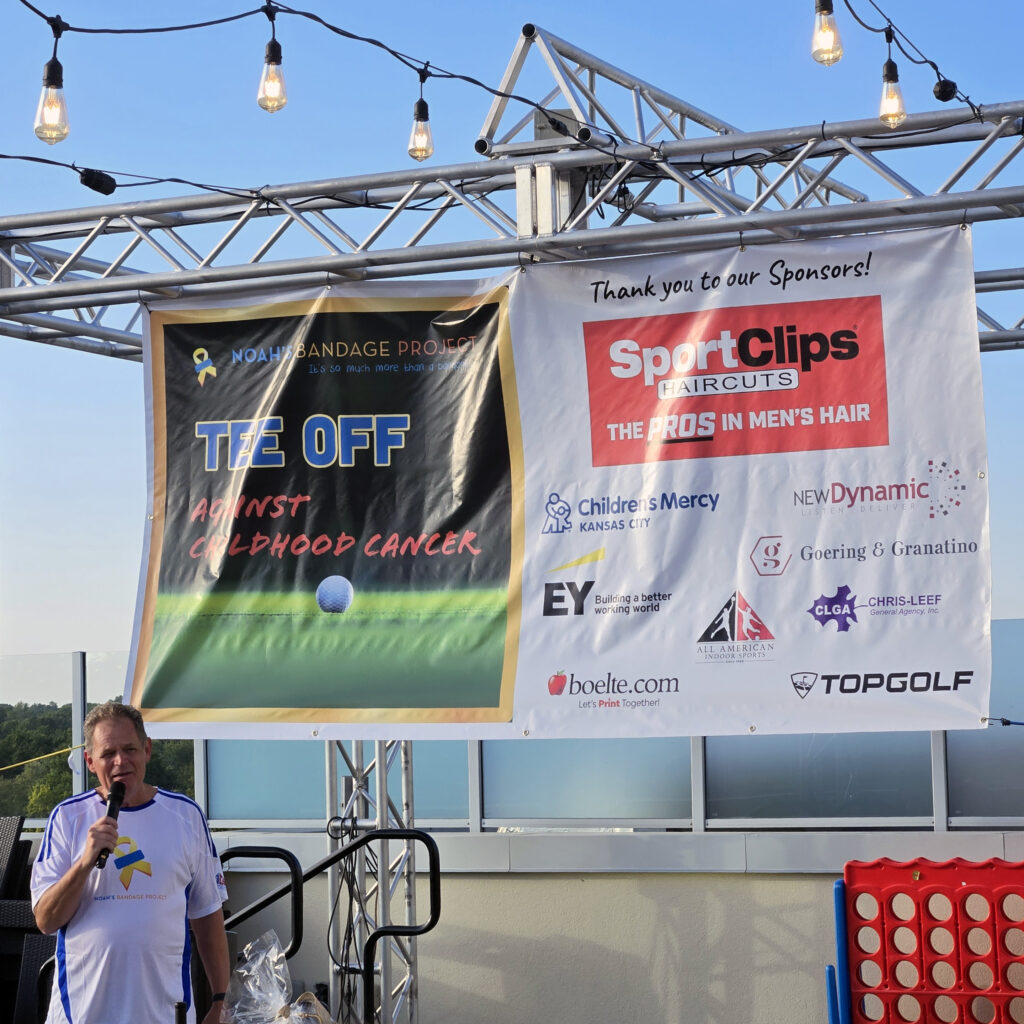Man in white shirt in the bottom left corner speaking into a microphone on a stage in front of two banners at the Tee Off Against Childhood Cancer event. The banner on the left is the promotional graphic for the event with the Noah's Bandage Project Logo and name of the event on top of an image of a golf ball on a golf course. The banner on the right features sponsor logos including New Dynamic's logos.
